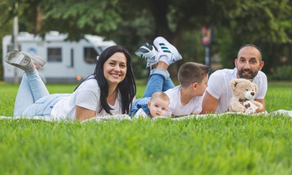 Happy family lying on grass with RV in the background, enjoying a sunny outdoor picnic.